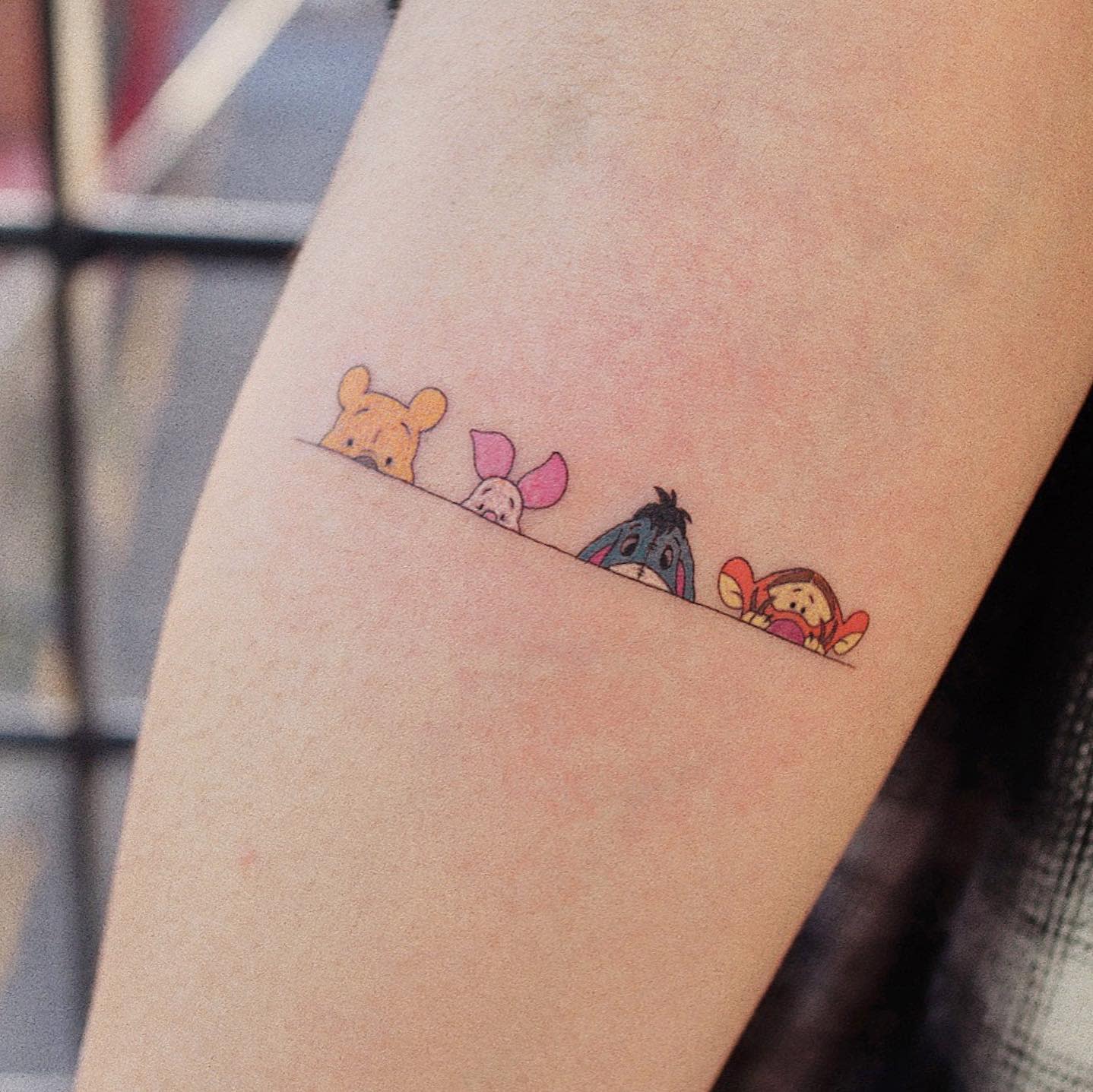 Colorful tattoo of Winnie the Pooh, Piglet, Eeyore, and Tigger peeking over a line on a person's arm